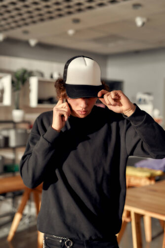 Be your own label. Stylish guy looking down while posing in custom apparel, black baseball cap and sweatshirt. Young man working at custom T-shirt, clothing printing company